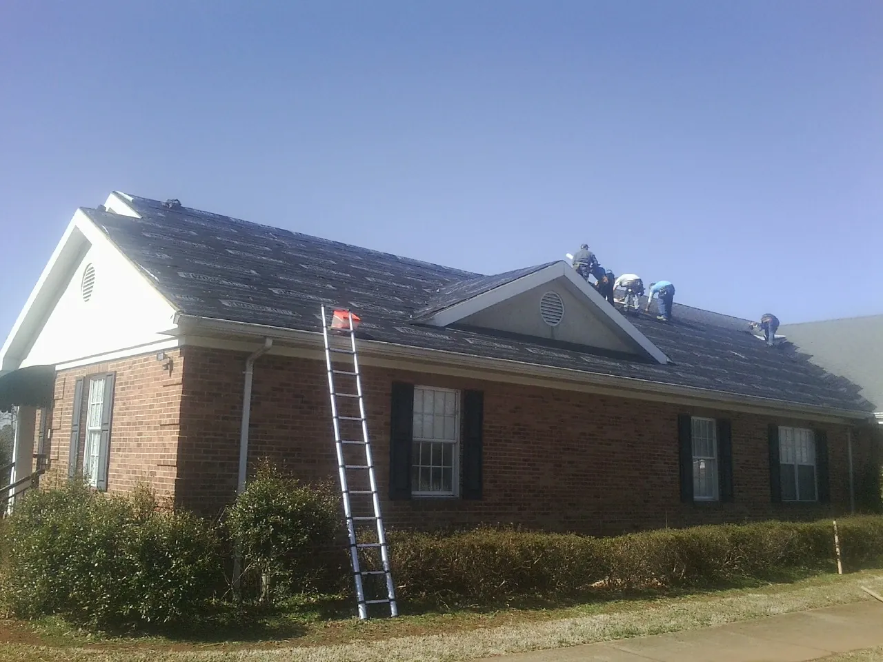 Crew tearing shingles off a brick ranch roof with a ladder set — Emergency Roof Repair work by our crew in Grand Bay