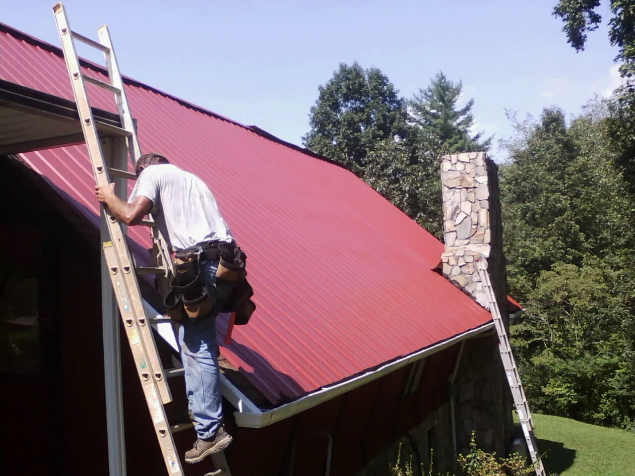 Roofer on a ladder beside a red metal roof and stone chimney — Soffit & Fascia Repair by Roofing Brothers in Grand Bay