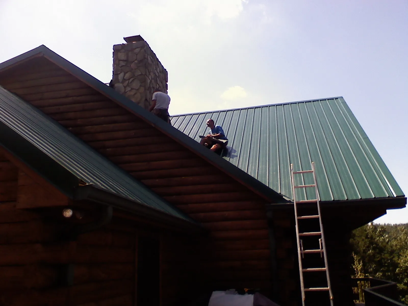 Roofers installing green metal panels beside a stone chimney on a log home in Grand Bay
